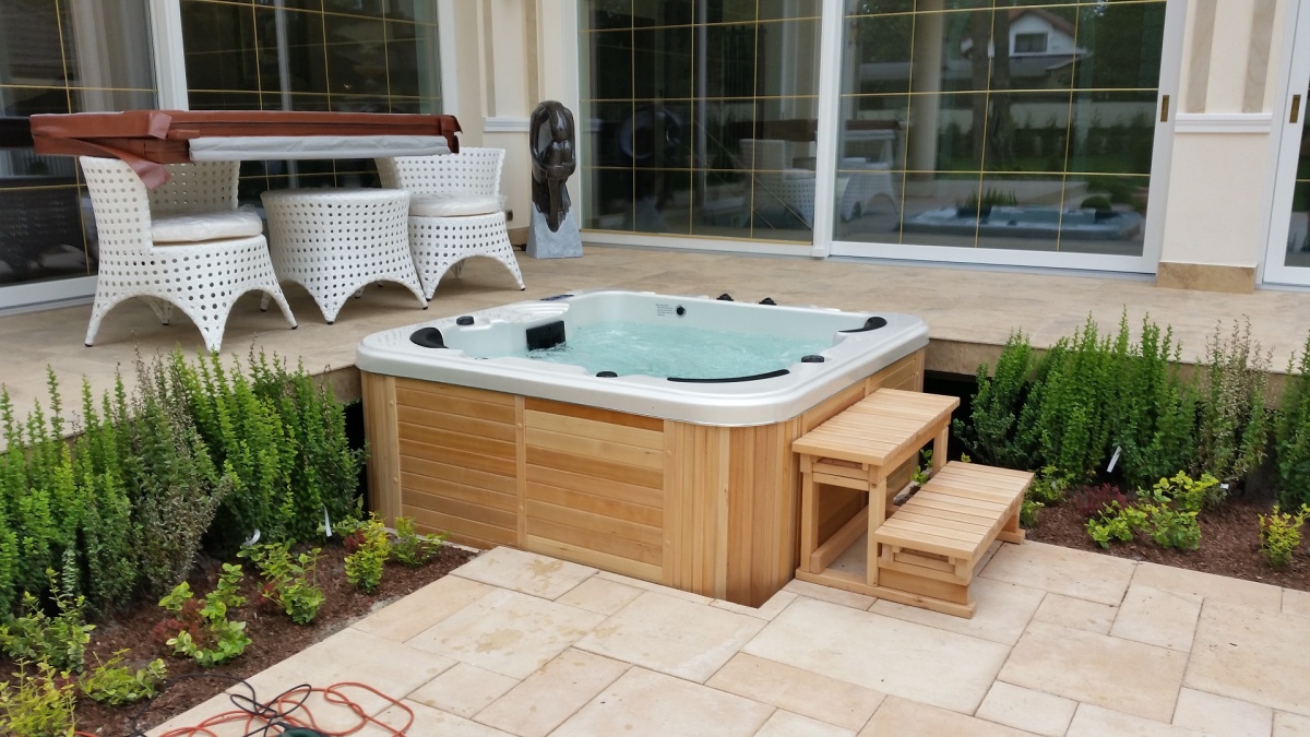 health by fun wooden hot tub on a tiled patio with small wooden steps, next to green shrubs and a seating area.