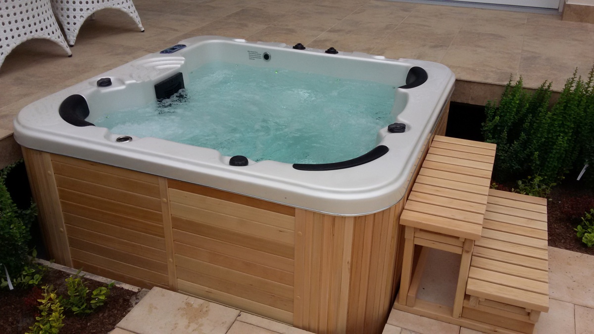 0 6 » Health By Fun health by fun outdoor wooden hot tub with bubbling water on a beige tiled patio, featuring black headrests and a wooden step ladder nearby.