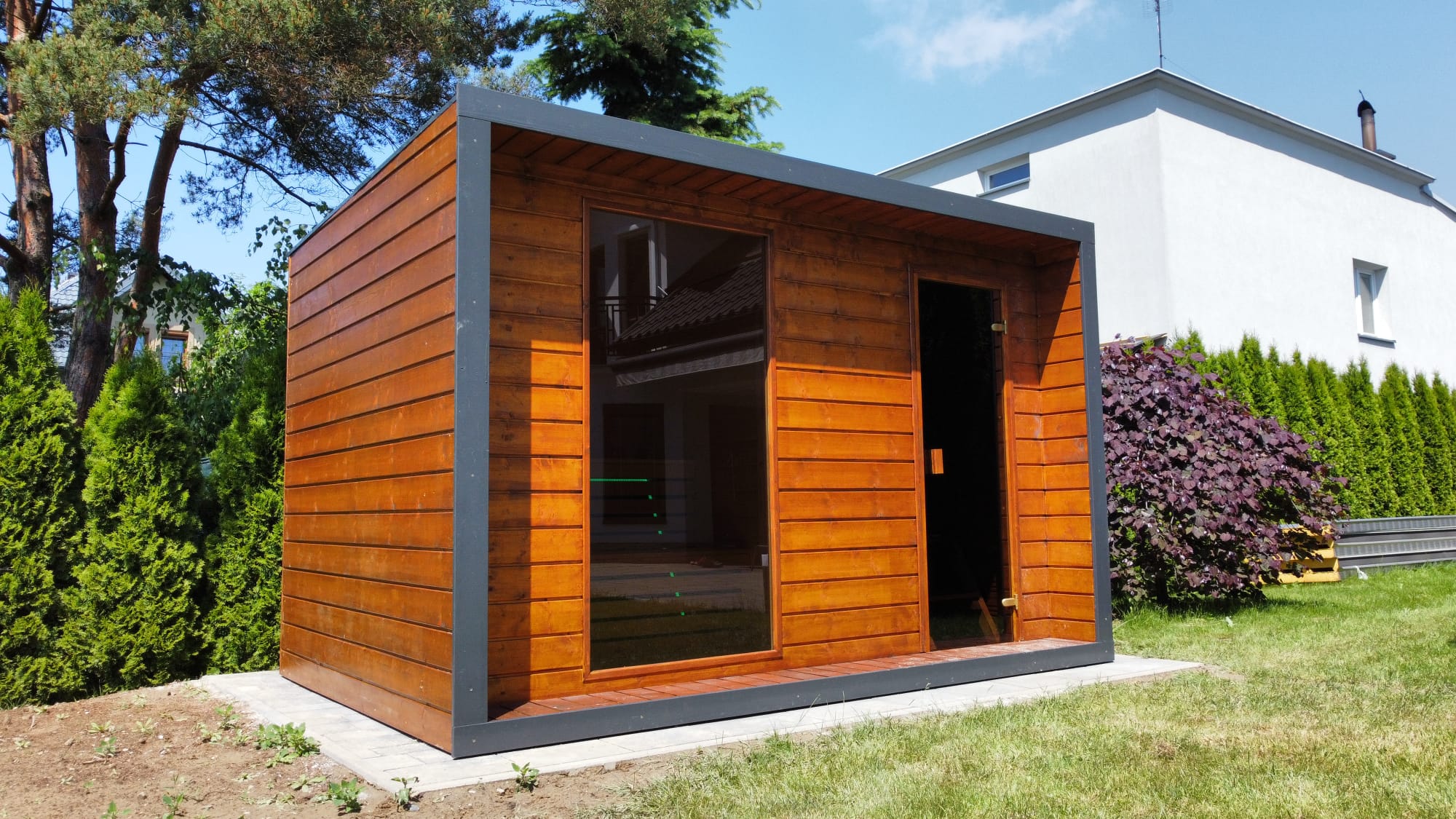 Modern wooden outdoor shed with a glass front and dark metal frame on a concrete pad, in a green backyard with trees and a white house behind it, sunny day.